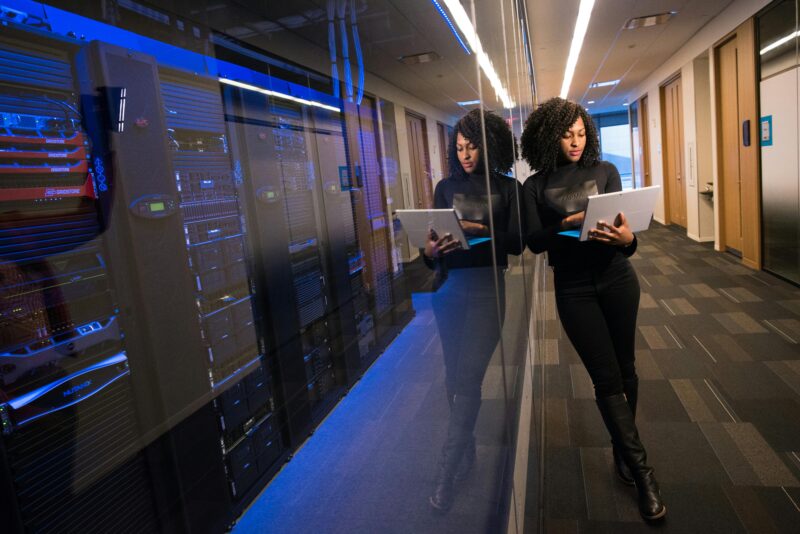 stock image of woman with laptop standing nexxt to server room in office building.